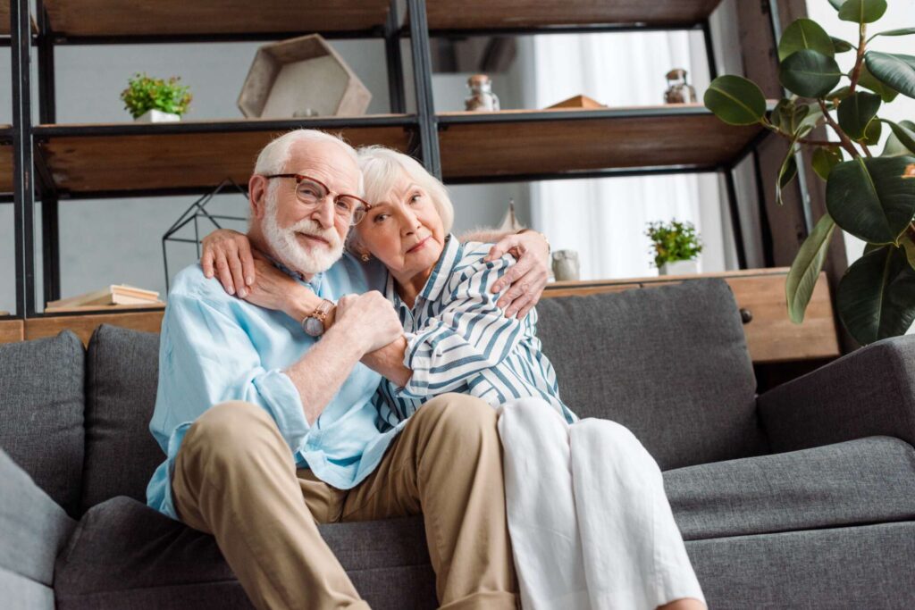 A white couple in their 70s sits on the sofa embracing each other.